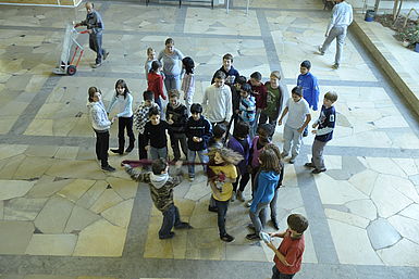 Schulhausszene: Junge reißt Mädchen Schal vom Hals, viele Jugendlichen stehen herum und schauen zu.
