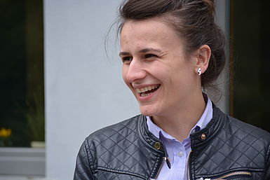 Lachende junge Frau mit dunkelbraunen Haaren, einer fliederfarbenen Bluse und einer schwarzen Lederjacke.