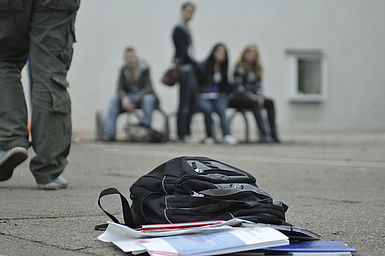 Rucksack liegt ausgekippt auf dem Boden, im Hintergrund sitzt eine Gruppe Jugendlicher.