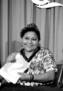 Rigoberta Menchú Tum sitzt auf einem Stuhl und hält ihre Auszeichnung zum Friedensnobelpreis in der Hand.